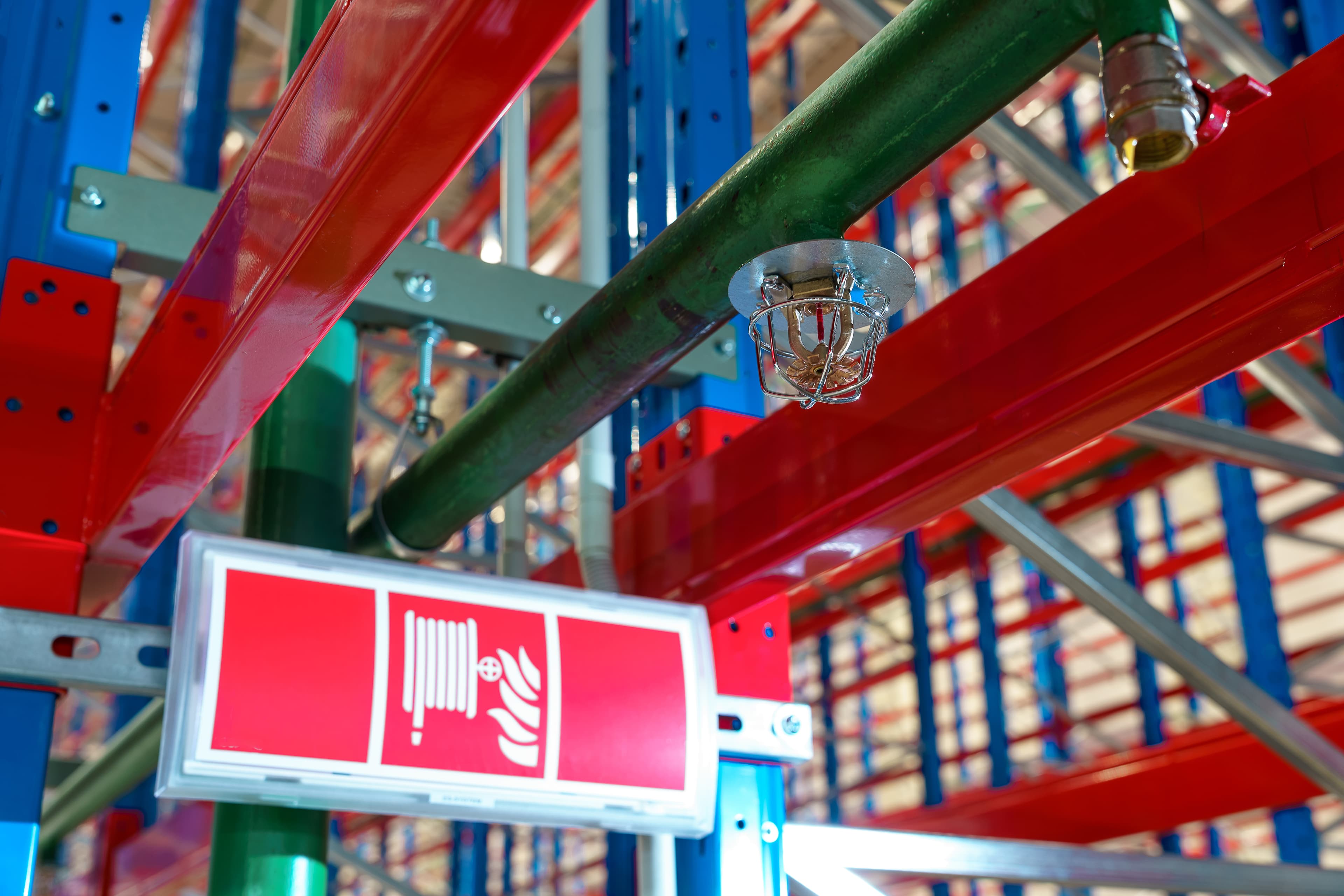 Red and blue warehouse racking with a fire sprinkler head on a green pipe overhead